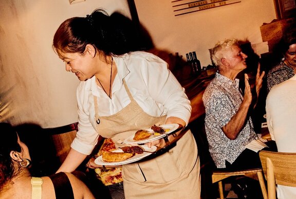A Peruvian waitress wearing a white shirt and beige apron at Barra Restaurant, serving plates of Peruvian food to diners.