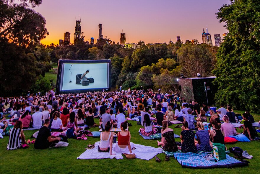 A crowd of people sitting on picnic blankets and watching a movie in an outdoor cinema surrounded by trees.