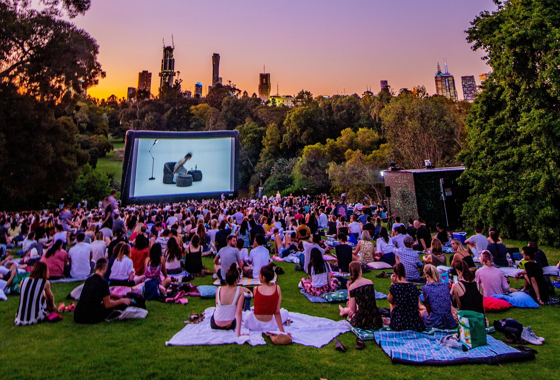 A crowd of people sitting on picnic blankets and watching a movie in an outdoor cinema surrounded by trees.
