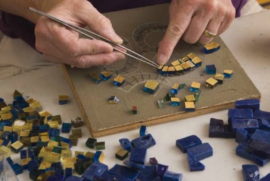 Person using a tool to place a mosaic on a tile, with other mosaic tiles to the side.