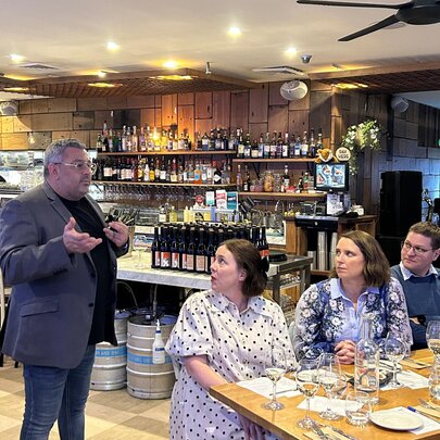 A room of people seated at tables in a restaurant, watching and listening to a standing person talking about wine.