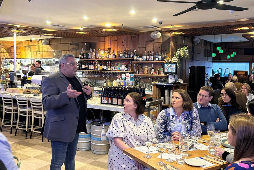 A room of people seated at tables in a restaurant, watching and listening to a standing person talking about wine.