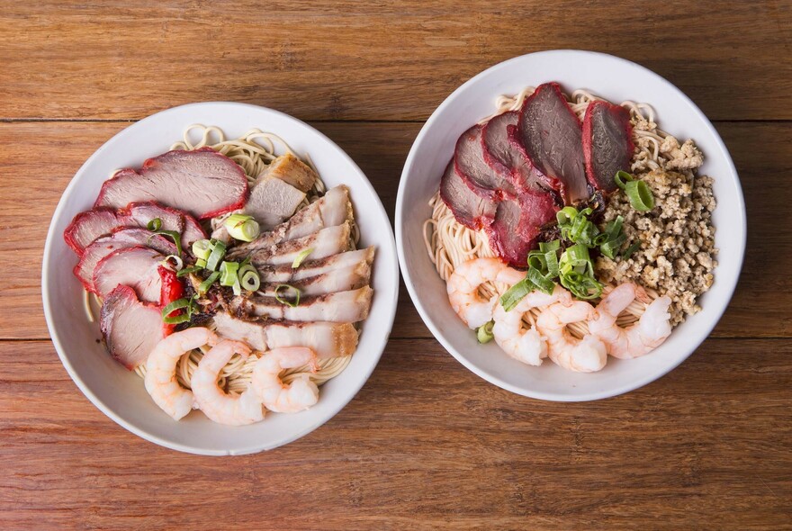 Wooden table with two bowls of noodle dishes with prawns and pork.