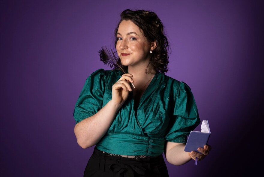 Woman with shoulder length hair, smiling at the camera and holding a feather to her face and a tiny purple book in her left hand.