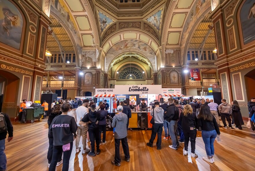 A crowd of people lining up in front of a coffee stall inside a large historic building with a dome ceiling.