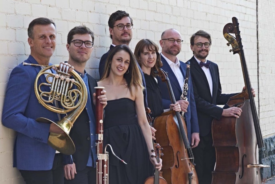Seven people from an ensemble lined up against a white brick wall and smiling while holding instruments. 