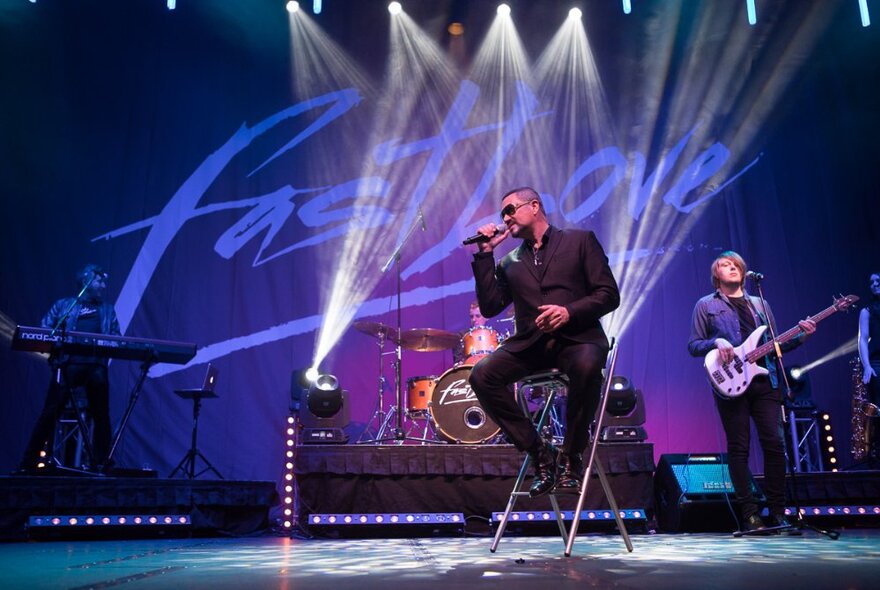 George Michael tribute performer seated on stage singing into microphone live with purple lighting and other band members.