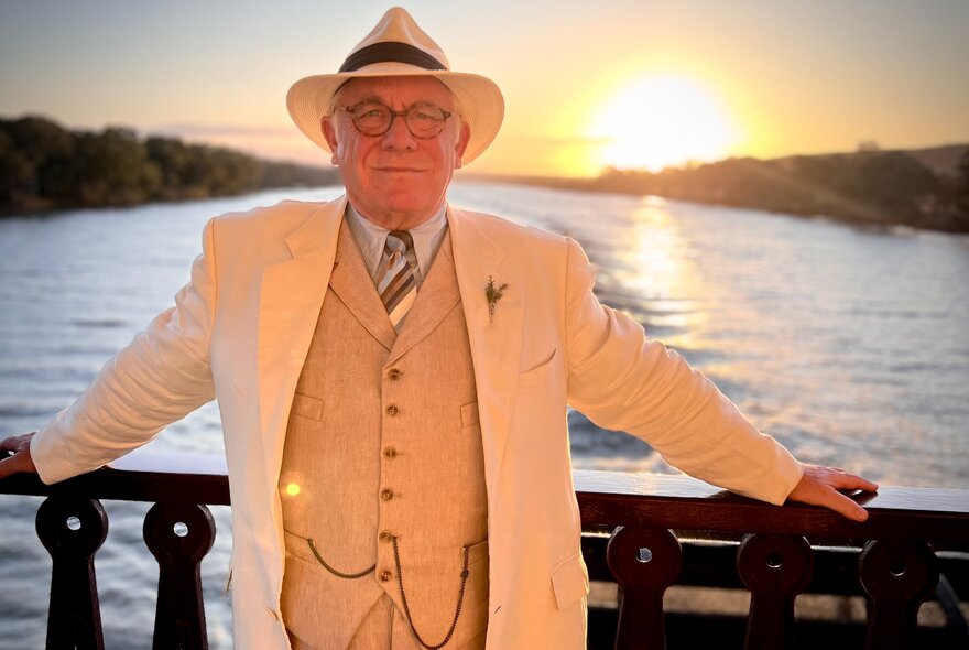 An older gentleman dressed in 1920s attire, leaning against the iron railing of a bridge over a river; sun setting behind him.