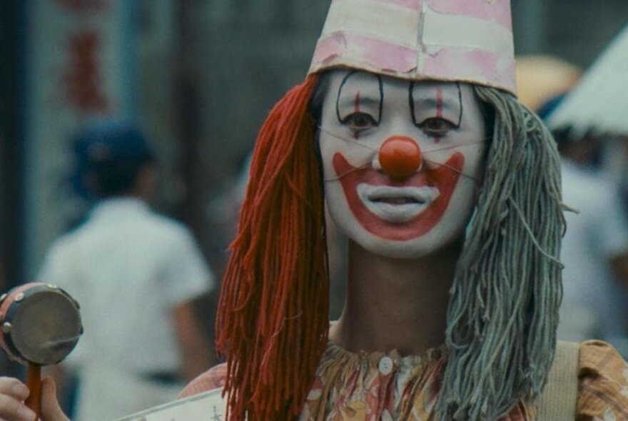 A still image from a Korean film showing a person dressed as a clown with wool for hair and a red nose. 