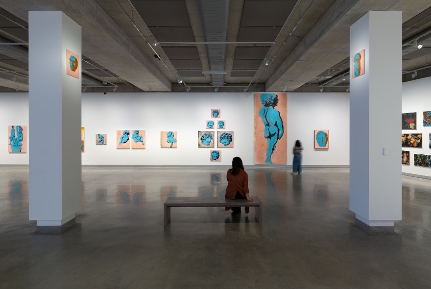 Person seated on a central bench in a large gallery space with white pillars and blue artworks on a long white wall.