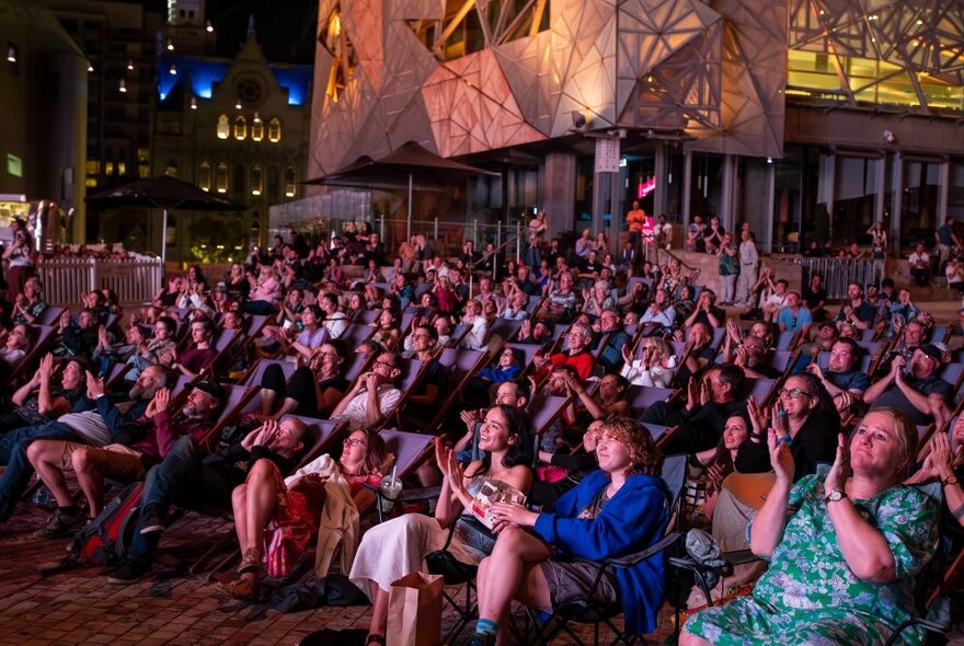 A seated audience in deck chairs at Fed Square at night.