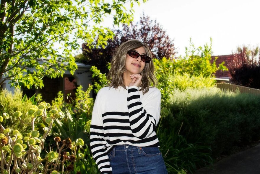 A woman looking happy wearing sunglasses and a striped jumper in a garden on a sunny day. 
