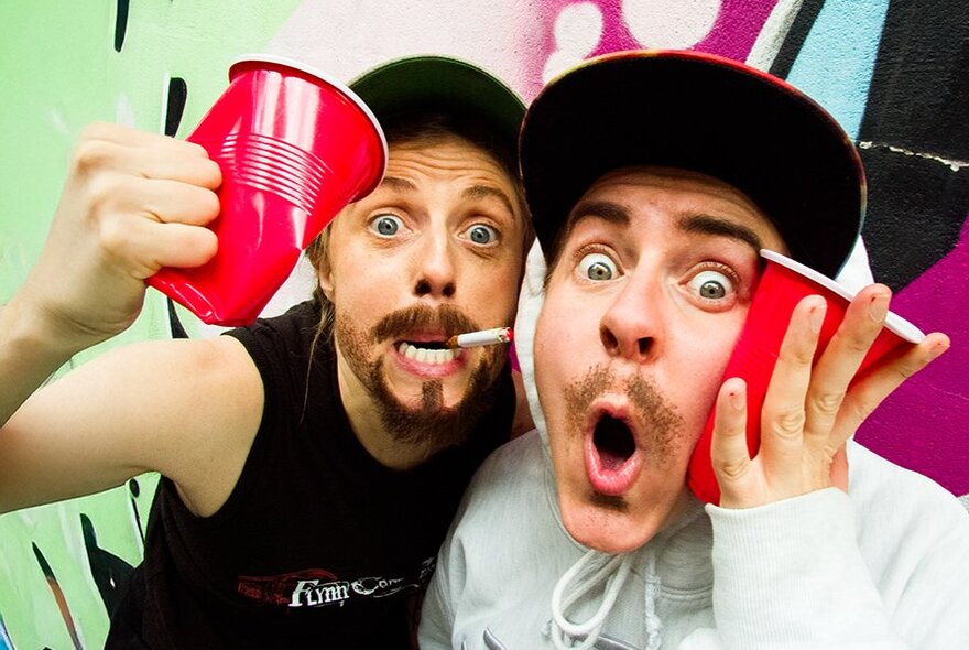 Two individuals posing with red cups, both wearing caps, with exaggerated facial expressions, as if having fun at a party.