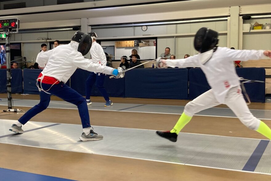 Two people dressed in white fencing outfits, fitted with head guards, fencing with swords on a fencing mat, indoors.