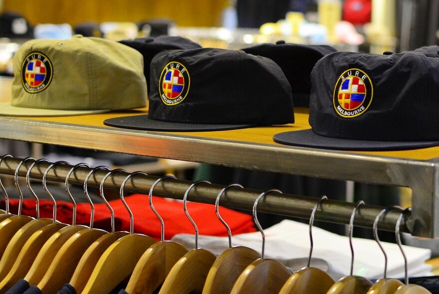 A row of three caps with logos on a shelf above a row of clothes hangers.