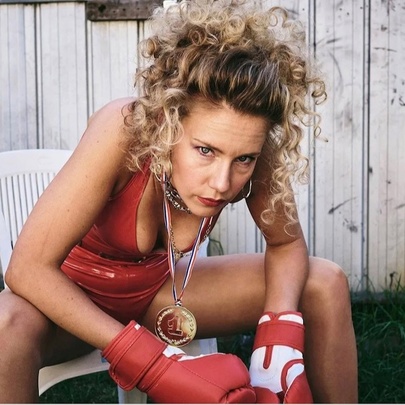 A portrait of a woman sitting forward in a white outdoor chair, wearing a red leather swimming costume and boxing gloves, a gold medal hanging around her neck. 
