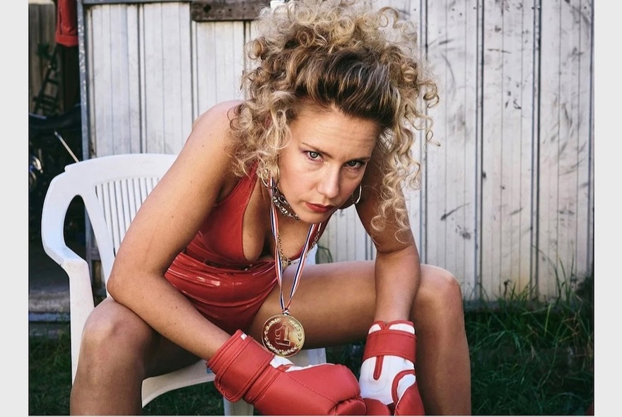 A portrait of a woman sitting forward in a white outdoor chair, wearing a red leather swimming costume and boxing gloves, a gold medal hanging around her neck. 
