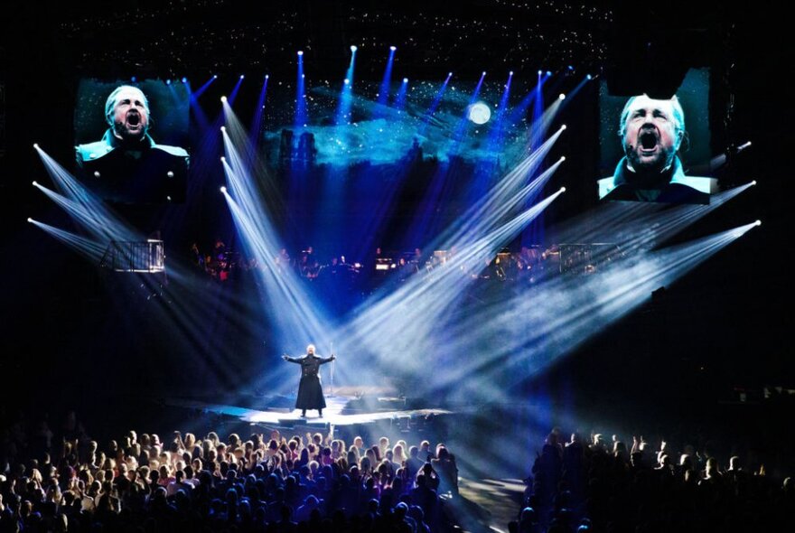 A solo male performer singing in a spotlight on stage in the musical Les Misérables The Arena Spectacular, with large screens at the rear of the stage projecting his face to the arena's audience.