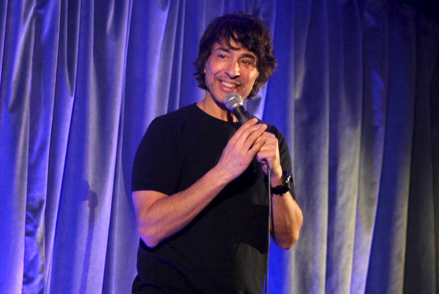 Comedian Arj Barker performing stand-up comedy live on stage in front of a blue curtain.