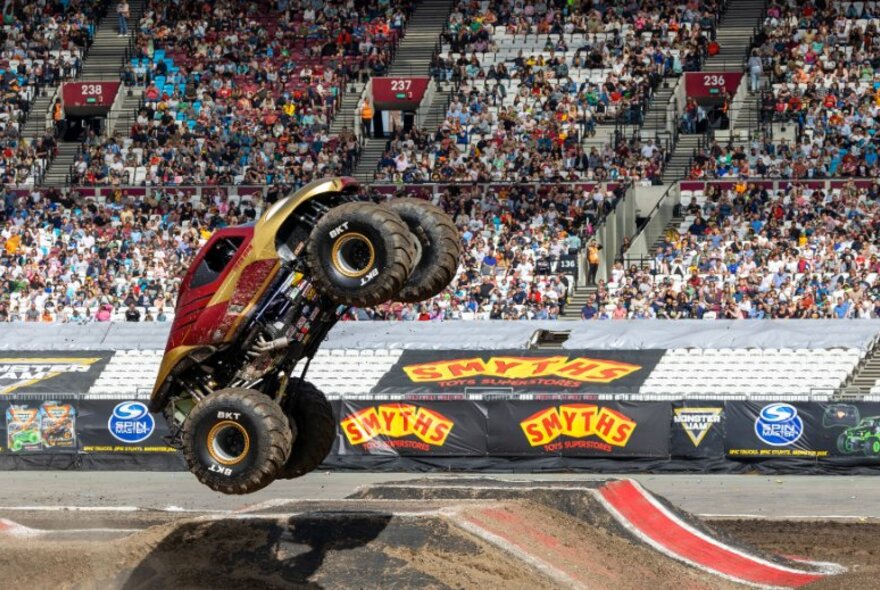 A monster truck flips in the air with wheels off the ground in an arena with a crowd looking on.