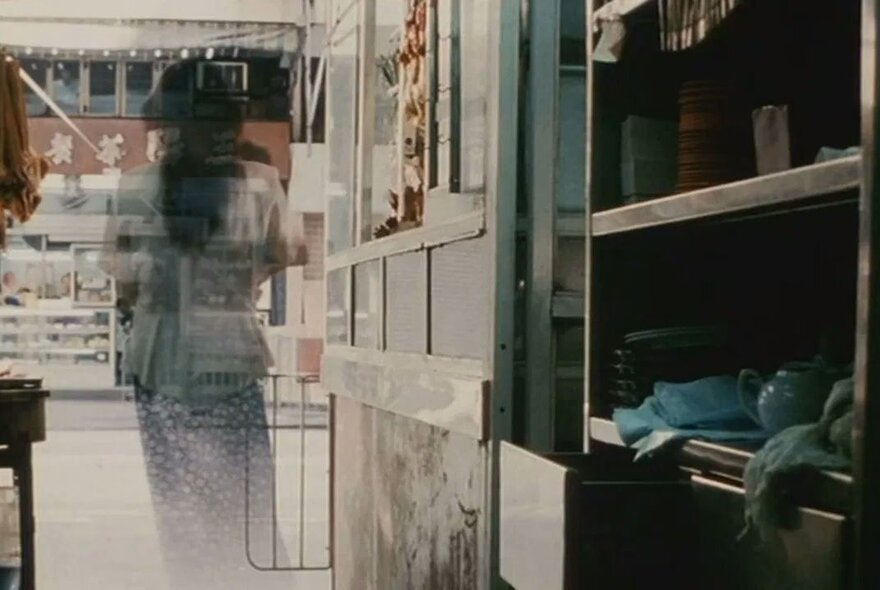 Still from the 2002 Hong Kong horror film The Eye of a blurry, transparent figure, appearing as a ghost, standing in a shop or room with shelves and counters.