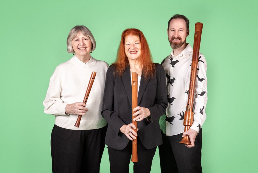 Three musicians smiling against a green background, each holding a wooden recorder of a different size.