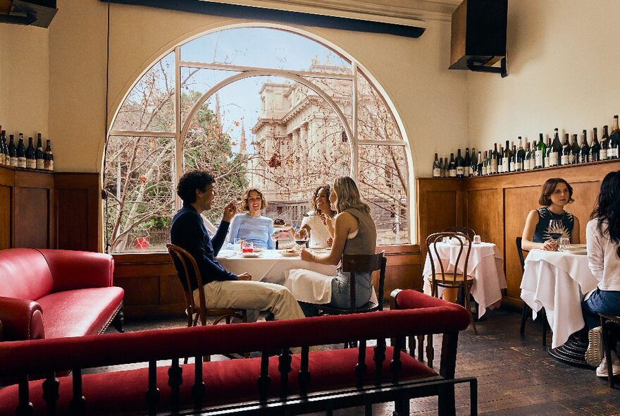 The interior of Kafeneion restaurant with diners seated at a table in front of a large arched window, in the foreground large red leather couches.