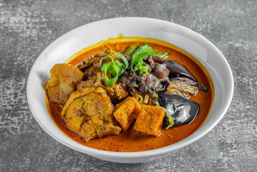 A white bowl of Asian soup in a red sauce with tofu and vegetables.