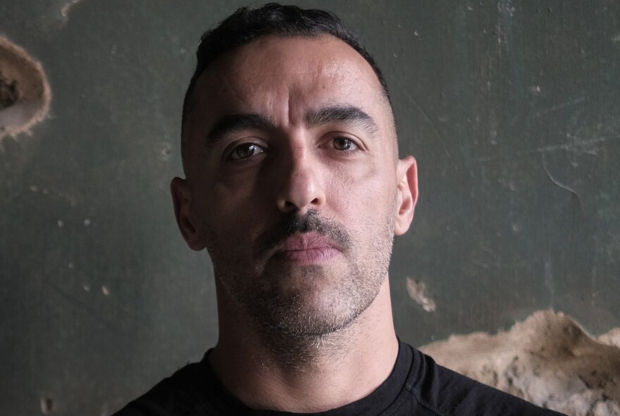 Portrait of journalist Antoun Issa, a man with short dark hair, a moustache, and a light stubble beard, looking directly forward against a dark, textured background.