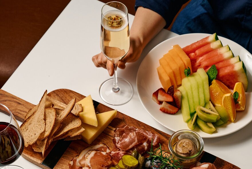 A woman's hand holding a champagne glass on a table, alongside a charcuterie board and a plate of sliced fruit.