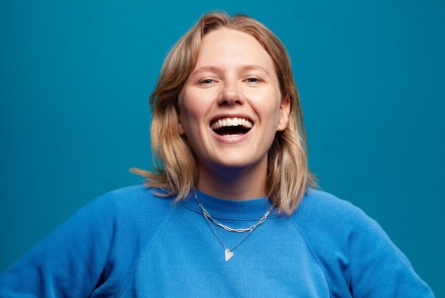 Laughing comedian with shoulder-length blonde hair against a blue background.