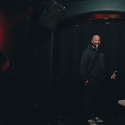 A dark image of a comedian on stage in a black hoodie and pants.
