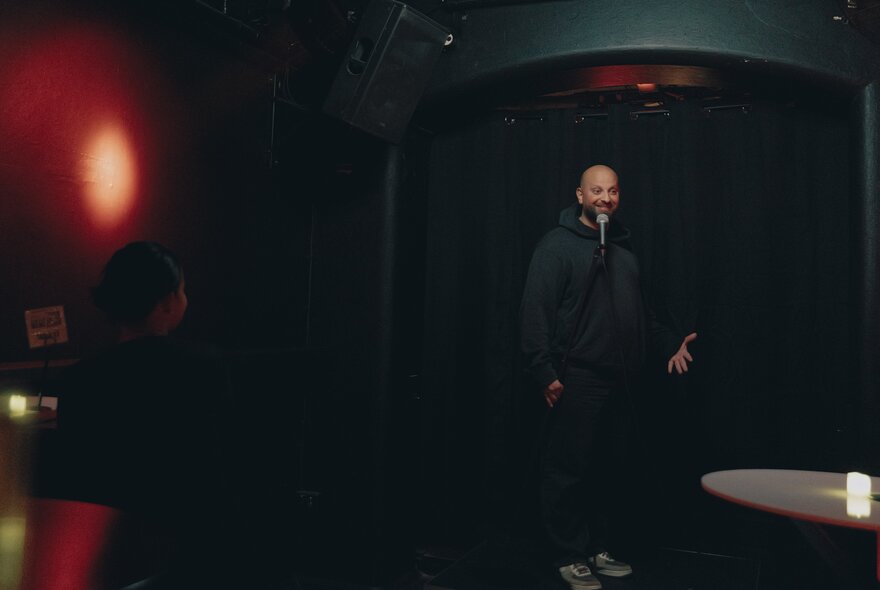 A dark image of a comedian on stage in a black hoodie and pants.