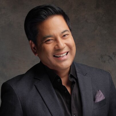 Singer Martin Nievera, a smiling middle-aged man with short, dark hair wearing a dark grey suit jacket over a black shirt posed against a textured grey background.