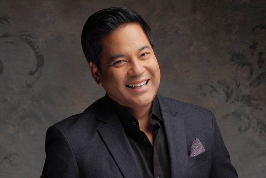 Singer Martin Nievera, a smiling middle-aged man with short, dark hair wearing a dark grey suit jacket over a black shirt posed against a textured grey background.