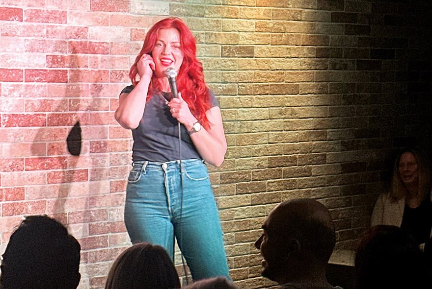 A stand-up comedian with long red hair, holding a microphone in her hand and talking on stage in front of an audience.