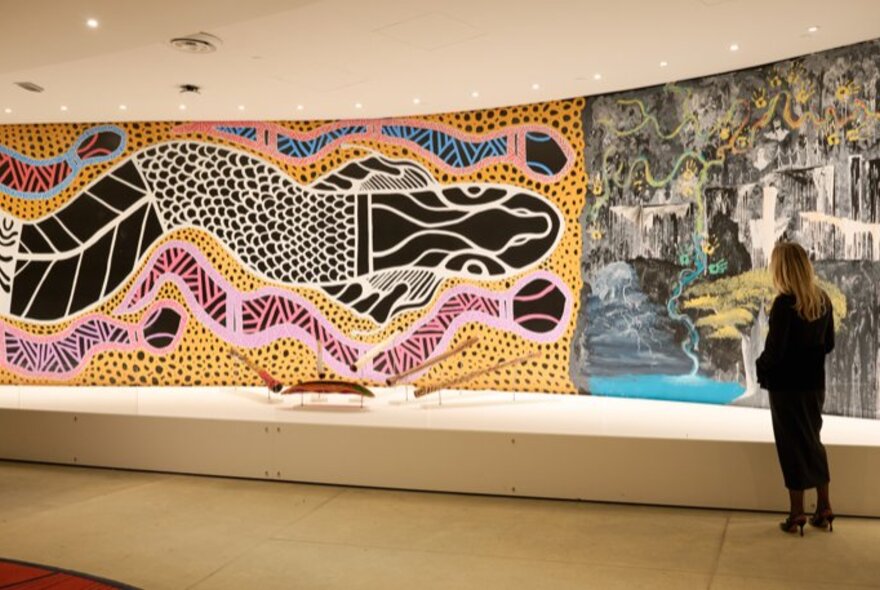 A gallery space featuring an indigenous mural artwork on the wall, a woman in black looking on.