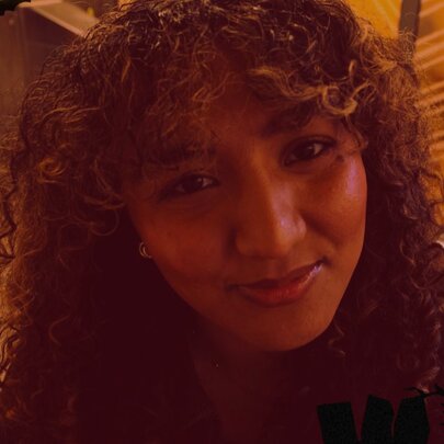 A young woman with dark, curly hair and a fringe, smiling slightly with warm lighting.
