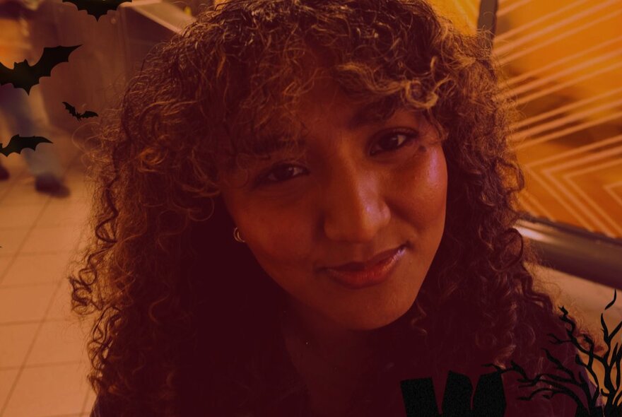 A young woman with dark, curly hair and a fringe, smiling slightly with warm lighting.