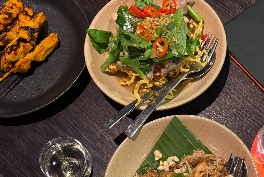 Dishes of Asian food with cutlery and glass on a table.