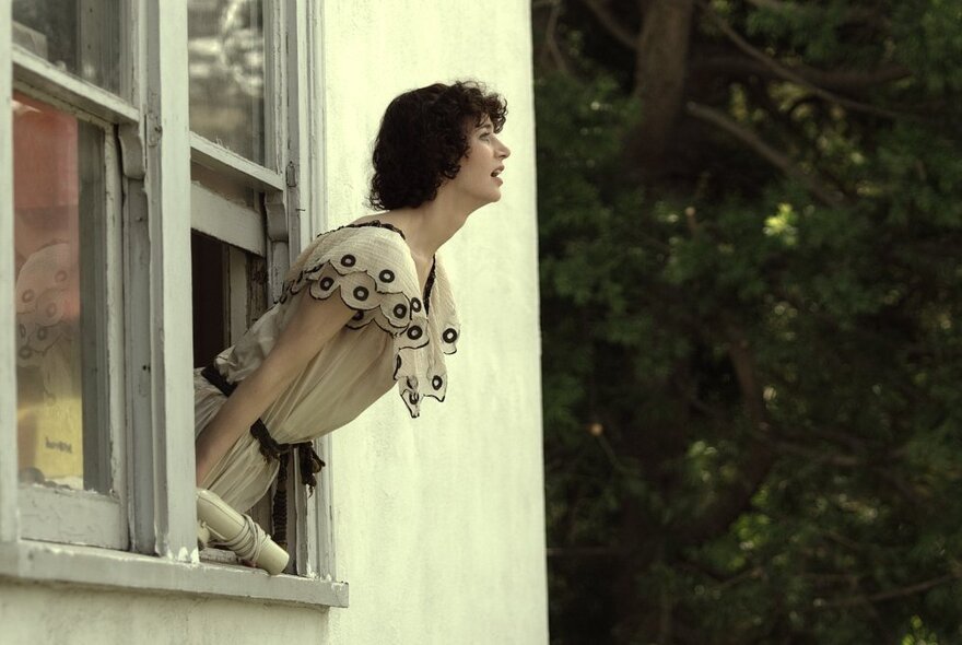 Film still of a woman in profile leaning out of a window of a house, green trees in the background.