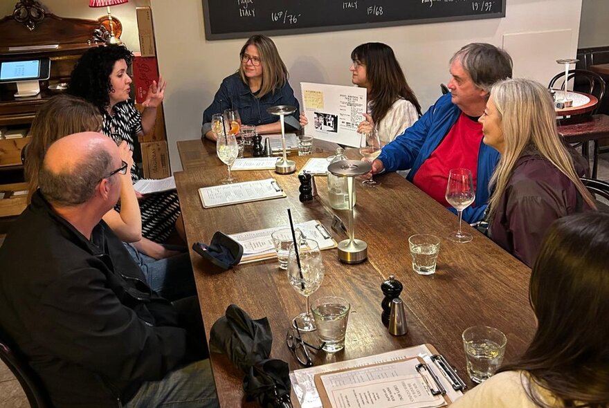 A group of people seated around a table in a restaurant, listening to someone talk, with menus and glasses of wine on the table.
