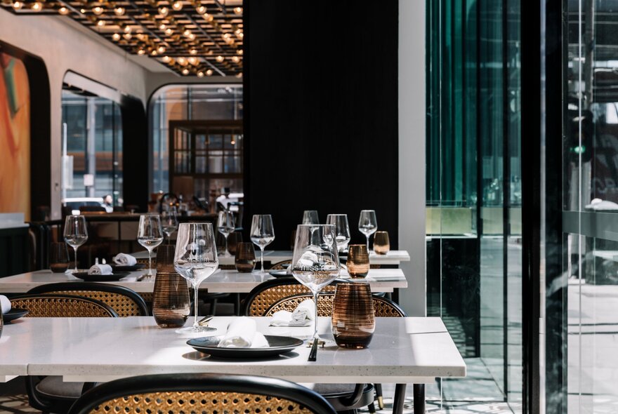 Interior of a hotel dining room with white tables set with wine glasses and plates, with large windows overlooking a city street. 