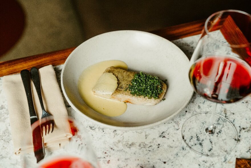 A fillet of fish with a sauce on a white plate, with a glass of wine alongside, on a restaurant dining table.