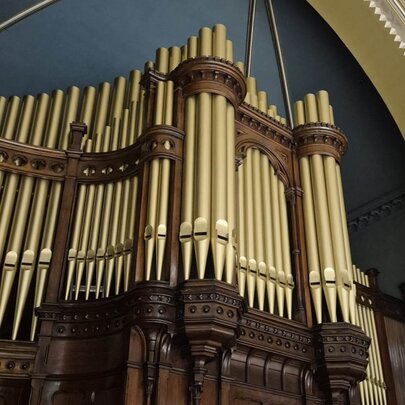 The grand pipe organ inside St Michael's Uniting Church in Melbourne, features gold-colored vertical pipes arranged in columns within a dark carved wooden frame under a dark blue vaulted ceiling.
