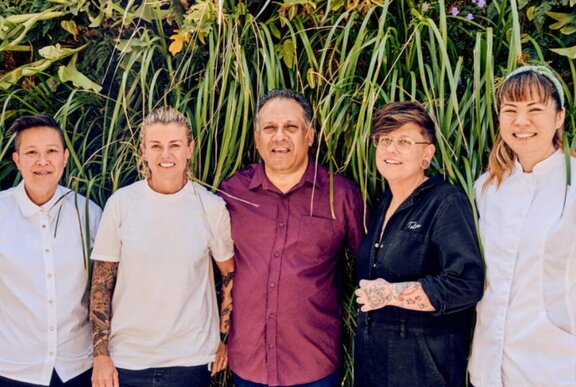 A row of chefs wearing uniforms standing against foliage.