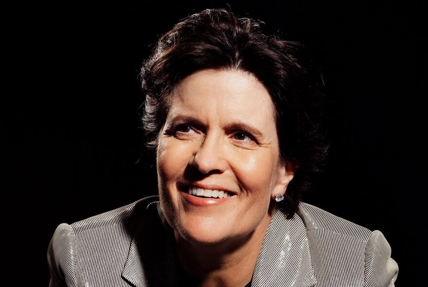 Journalist Kara Swisher smiling against a black background, wearing a shiny striped jacket.