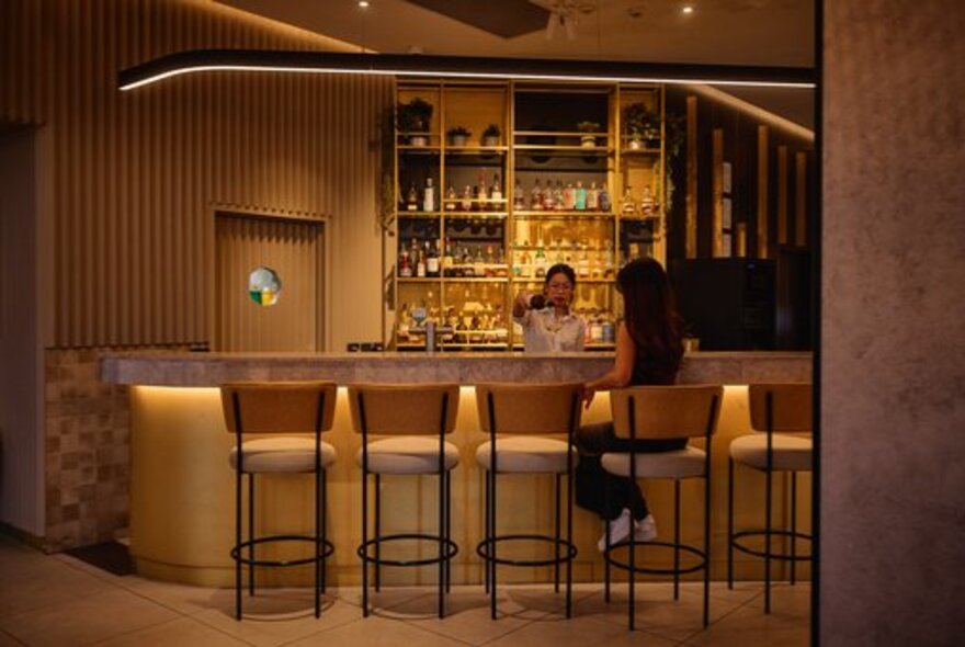 The bar at One Queen restaurant, with a patron seated on a high stool and a bartender preparing drinks, with warm lighting.