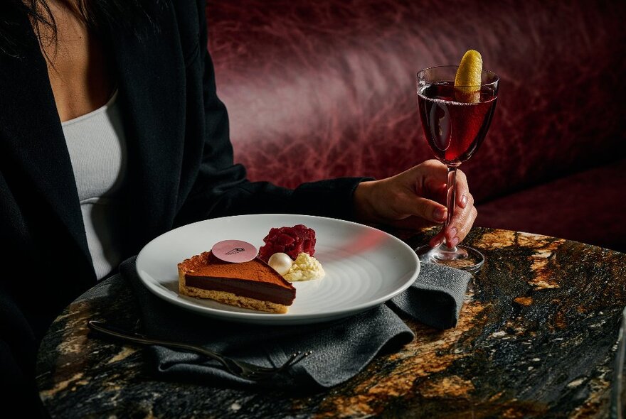 A woman wearing a black blazer seated at a burgundy booth and marble table, with a cocktail in her hand and a plate with a decadent chocolate tart in front of her.