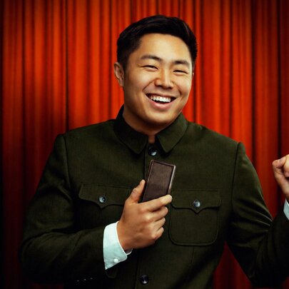 Comedian Nathan Chin on stage with an old-fashioned microphone, in front of a red stage curtain.